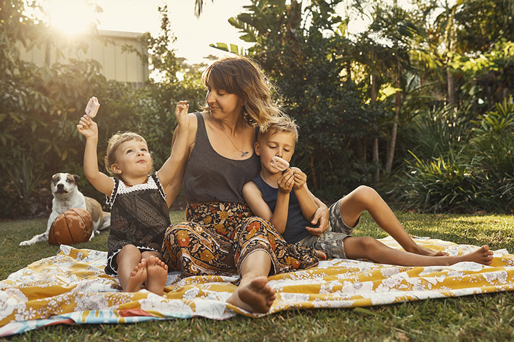 Mother sitting on a picnic blanket with two young children enjoying ice creams in a backyard garden with a dog nearby